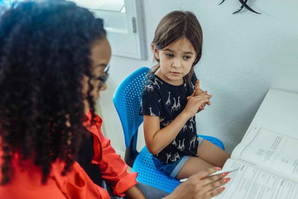 High angle of ethnic female teacher with exercise book speaking with pupil during lesson at home
